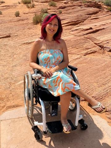 Photo caption: Kam Redlawsk in her wheelchair. Kam has pink streaks in her dark hair and she is wearing a strapless blue and orange dress. Photo courtesy of Kam Redlawsk.